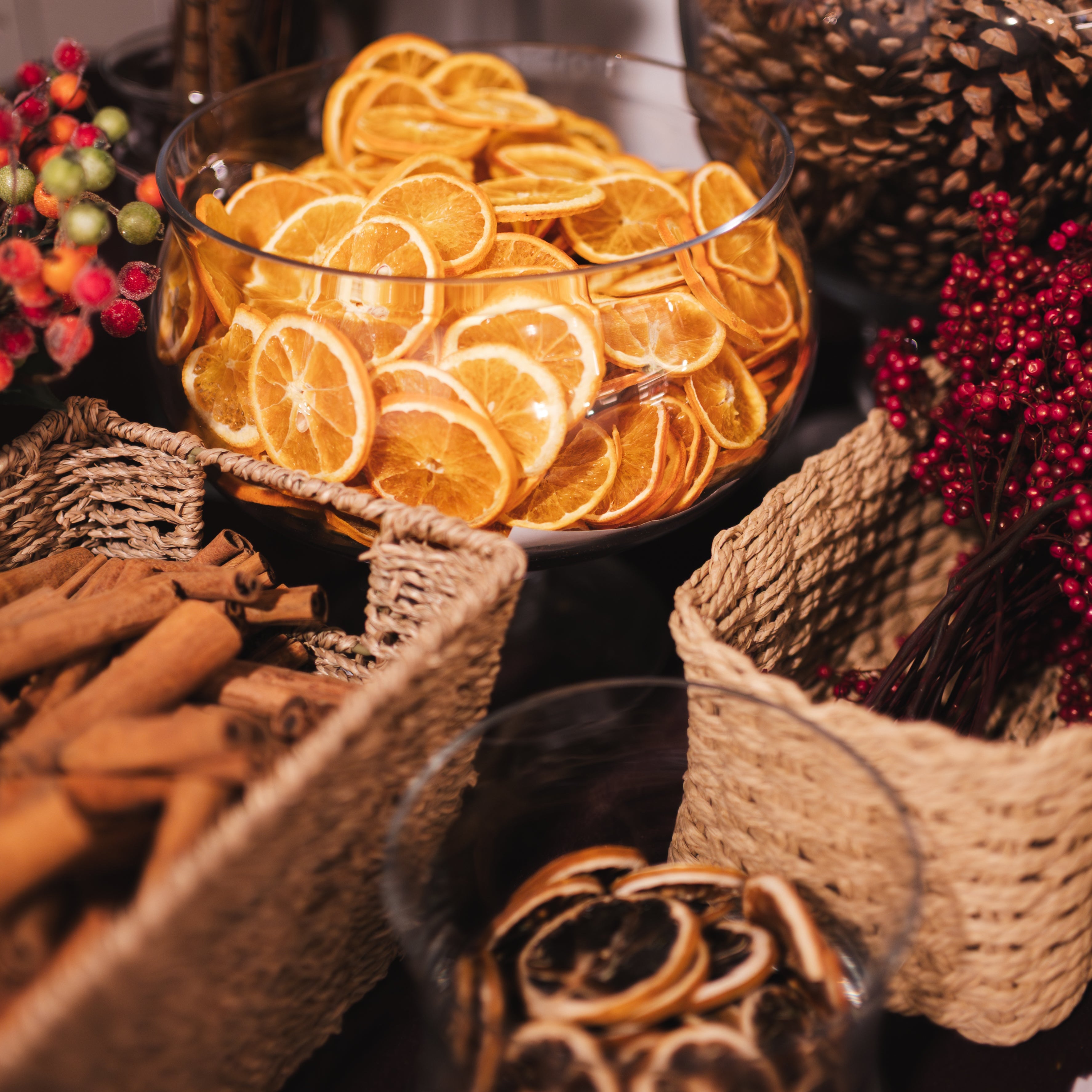 Dried orange slices arranged in clear glass bowls and woven baskets, with cinnamon sticks and red berries in a warm, rustic setting.