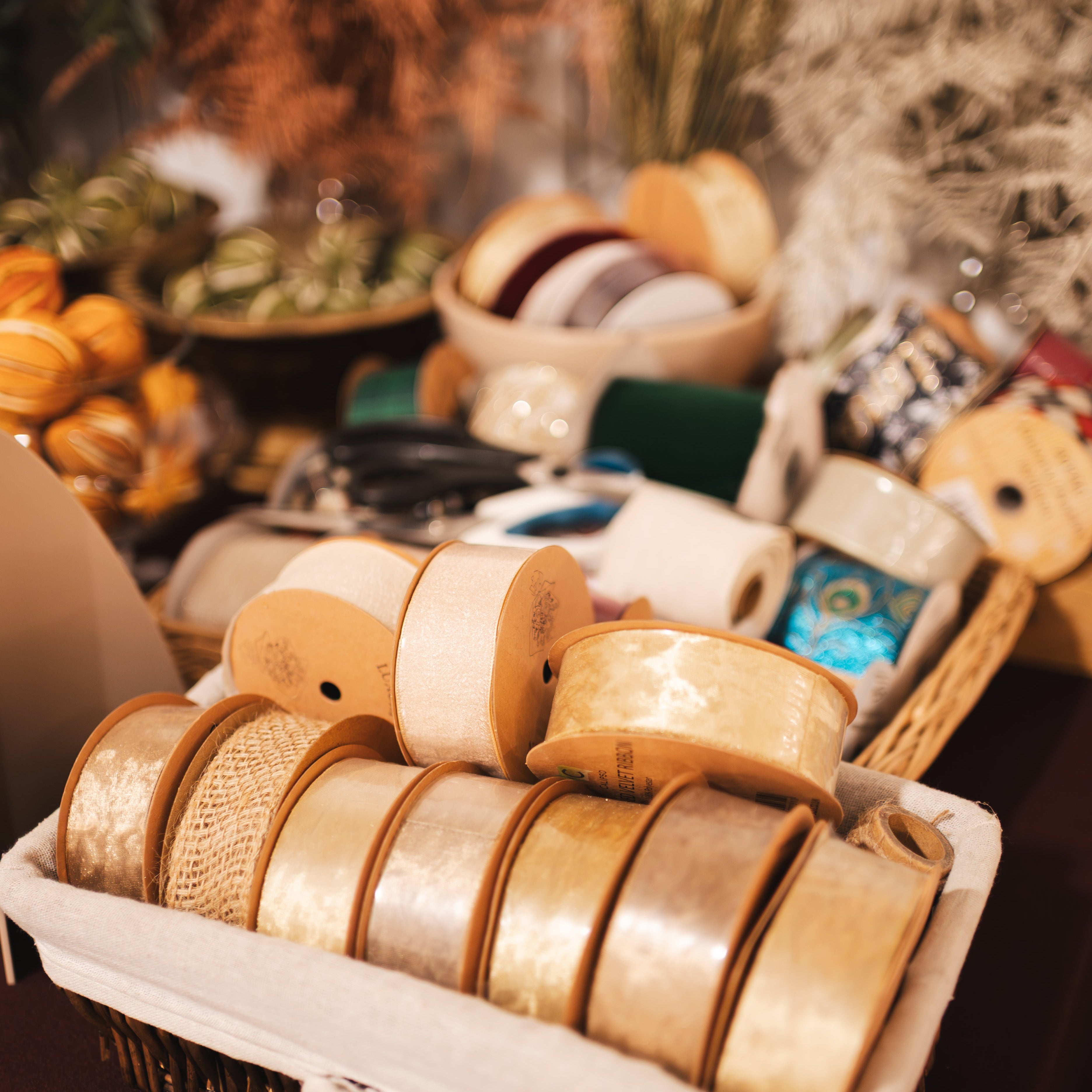 A wicker basket lined with cream fabric holds multiple rolls of ribbon in warm neutral and metallic tones, photographed in a softly styled indoor setting with other decorative items blurred in the background.