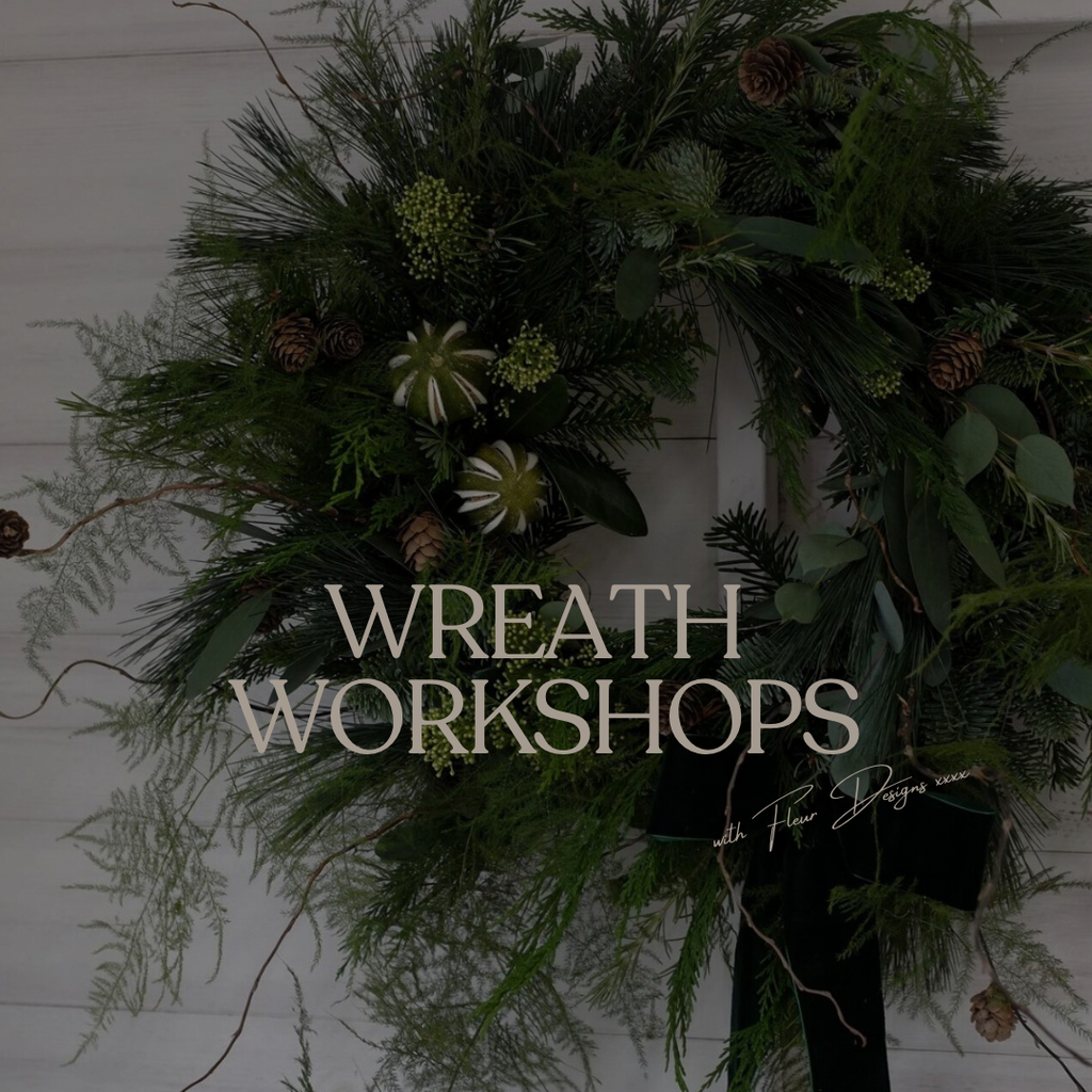 A dark green evergreen wreath with pinecones, green berries, and white starburst flowers, hung against a white paneled wall. Large text across the center reads 'WREATH WORKSHOPS' with a smaller signature-style line below.